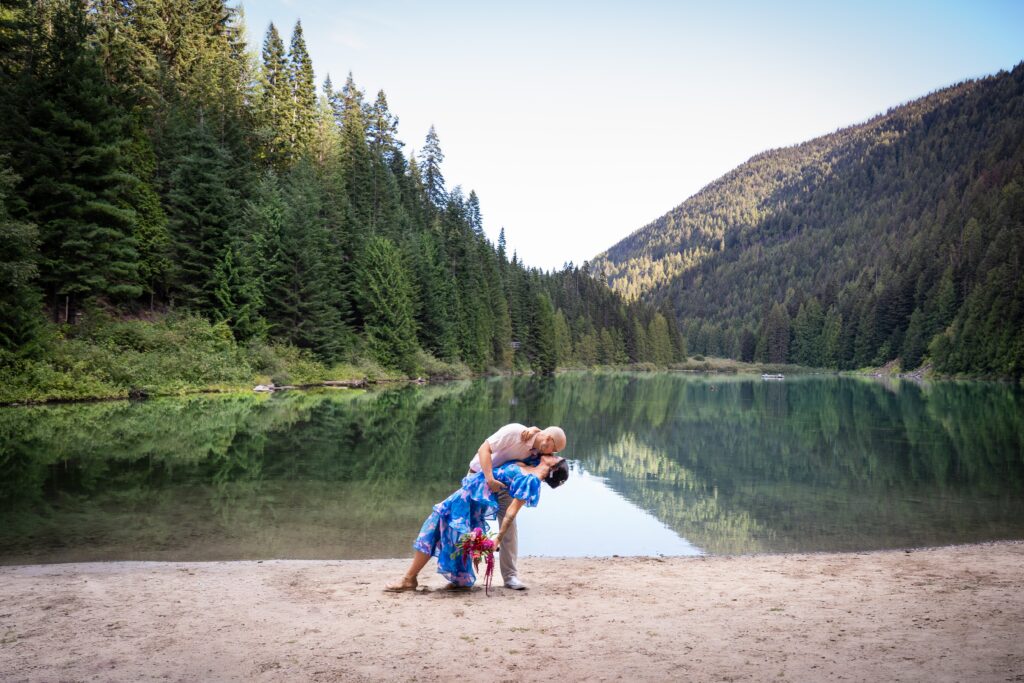 groom dips bride as they kiss in front of a lake with mountains